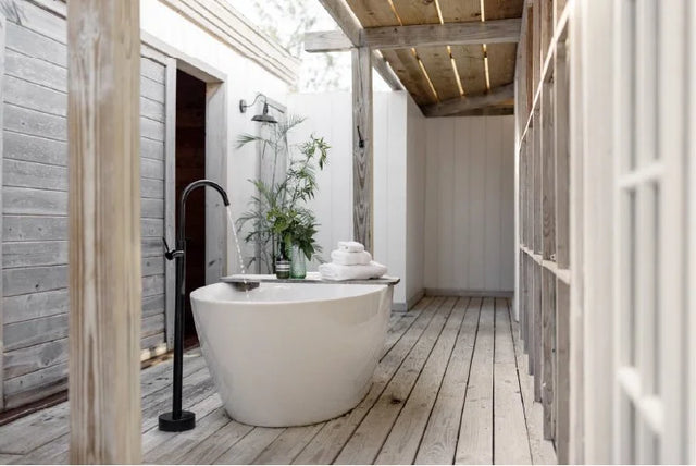 A modern freestanding oval bathtub in a rustic outdoor bathroom setting with light wood flooring, weathered wood walls, a black faucet, and a small plant arrangement.