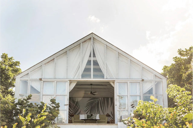 A large white event pavilion or greenhouse-style venue with a steep gable roof, draped white curtains, and a wide open central entrance revealing a decorated interior, surrounded by green trees and foliage.