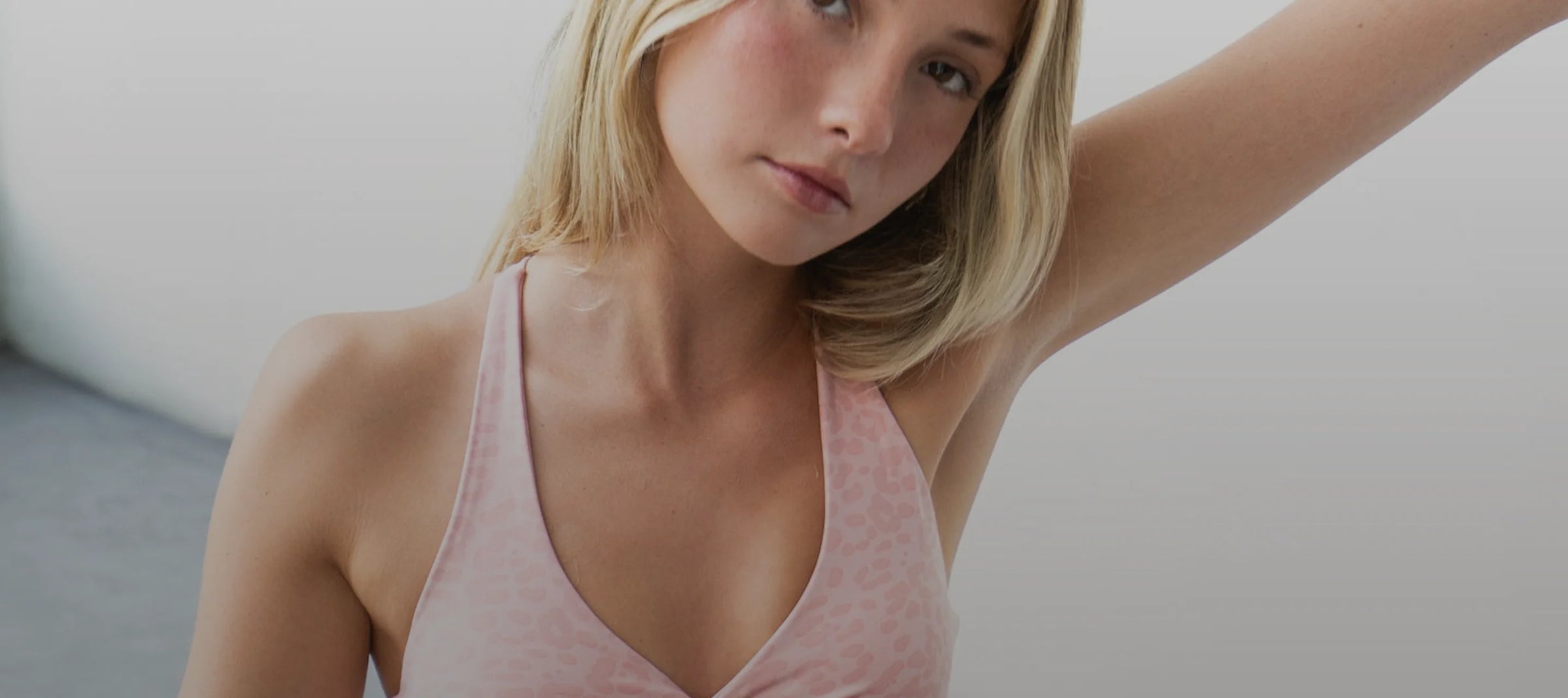 A blonde woman wearing a light pink halter-neck tank top, shown from the chest up against a soft gray background.